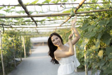 Beyaz elbise içinde gülümseyen, yemyeşil bahçede dans eden mutlu genç Asyalı kadın. Neşeli kadın doğada güneşli bir günde açık hava bitki çardağının keyfini çıkarıyor.