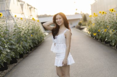 Ay çiçeği bahçesinde yaz akşamının tadını çıkaran güzel mutlu Asyalı kadın. Gün batımında altın ışıkla gülümseyen genç yetişkin portresi