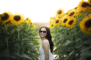 Ayçiçeği tarlasında gülümseyen beyaz elbiseli ve güneş gözlüklü mutlu bir kadın. Neşeli kadın yaz güneşinin tadını çıkarıyor ve sıcak hava tatili boyunca doğa.