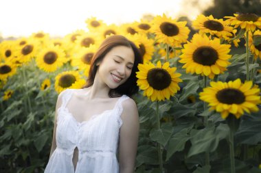 Güzel mutlu Asyalı kadın huzurlu gülümsemesiyle canlı ayçiçeği tarlasında yaz günbatımının tadını çıkarıyor. Doğada altın güneş ışığı altında huzurlu bir an.
