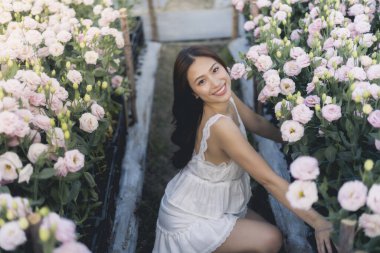 Pembe çiçek bahçesinde gülümseyen güzel Asyalı kadın. Çekici dişi seranın içine çömelmiş çiçek açan çiçeklerle çevrili, doğadaki huzurlu anın tadını çıkarıyor.