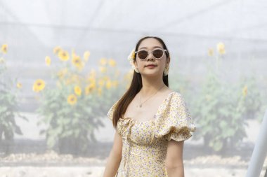 Güzel sarı elbiseli, güneş gözlüklü, kendine güvenen Asyalı kadın çiçek bahçesinde huzurlu bir yaz gününün tadını çıkarıyor. Doğadaki barışın portresi.