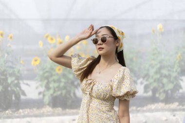 Çiçekli elbiseli ve güneş gözlüklü şık Asyalı genç kadın güzel ayçiçeği tarlasında sakin bir yaz gününün tadını çıkarıyor. Doğada sakin moda portresi