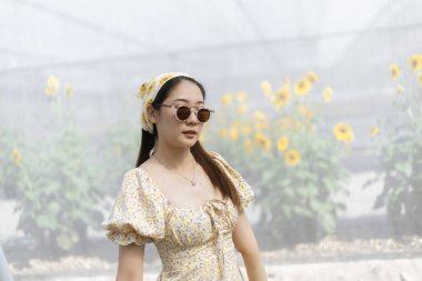 Yazlık elbise ve güneş gözlüğü giymiş zarif Asyalı kadın güzel bir ayçiçeği tarlasında huzurlu görünüyor. Doğanın ve güneş ışığının tadını çıkaran bir insan portresi.