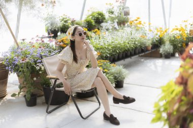 Güneşli serada sandalyesinde dinlenen sakin bir kadın. Bu yaşam tarzı onu güzel çiçek ve bitki düzenlemelerinin arasında otururken gösteriyor. Huzurlu ve mutlu hissediyor.