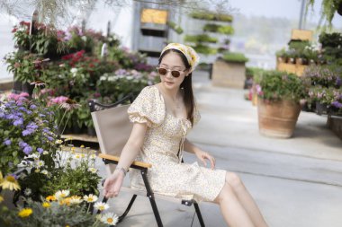 Şık, genç Asyalı kadın kendini huzurlu ve rahat hissediyor. Serada sandalyede oturuyor. Rengarenk çiçeklerle ve yeşil bitkilerle çevrili bahçedeki boş zamanların tadını çıkarıyor.