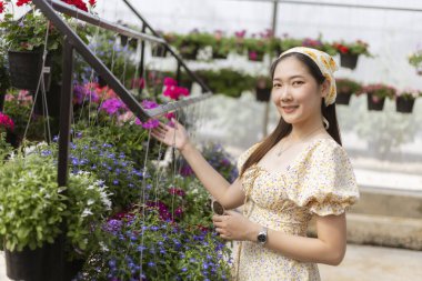 Mutlu gülümseyen Asyalı kadın çiçekçide bahçeyle eğleniyor. Seradaki renkli bitkilere dokunur. Hobisinin neşeli ve güzel bir sahnesidir.
