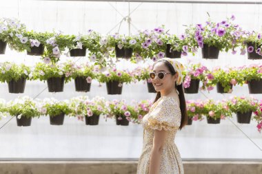 Bahçede neşeyle gülümseyen güneş gözlüklü mutlu Asyalı kadın. Arka planda bahar mevsiminde serada asılı petunya saksıları duvarı yer alıyor