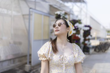 Güneş gözlüğü takan mutlu Asyalı kadın dışarıda yaz gününün tadını çıkarıyor. Seranın önündeki moda portresi, güneşli tatilde huzurlu ve rahat.