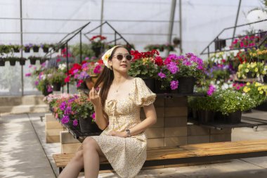Renkli çiçeklerle çevrili serada oturan şık elbiseli ve güneş gözlüklü mutlu genç Asyalı kadın. Neşeli insan, bitki yetiştirme odasında güzel bir günün tadını çıkarıyor.