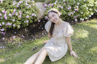 Parktaki yeşil çimlerin üzerinde oturan mutlu Asyalı kadın. Gülümseyen dişi, güneşli yaz gününde güzel çiçek bahçesinde portre oturumundan zevk alır.