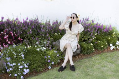 Huzurlu Asyalı kadın bahçede dinleniyor. Mor çiçeğin arasında oturan kadın elbise ve güneş gözlüğü takıyor, yaz günü açık havanın tadını çıkarıyor.