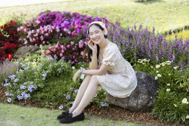 Renkli çiçek bahçesinde kayaların üzerinde otururken mutlu gülümseyen genç Asyalı güzel bir kadın. Huzurlu bir kadın, huzurlu bir günün tadını çıkarıyor. Doğada dinleniyor.