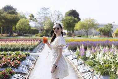 Beyaz elbiseli neşeli genç Asyalı kadın, güneşli bir günde elinde turuncu bir meyve ile tarlada renkli çiçeklerle güzel bir çiçek bahçesinde gülümsüyor.