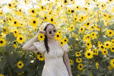 Güzel bir ayçiçeği tarlasında barış işareti ile poz veren şık elbiseli ve güneş gözlüklü mutlu genç Asyalı kadın. Neşeli, kaygısız açık havada yaz günü portresi