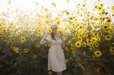 Yazın ayçiçeği tarlasında duran beyaz elbiseli güzel mutlu kadın. Güneşli bir günde doğanın tadını çıkaran genç bir kadının huzurlu portresi.