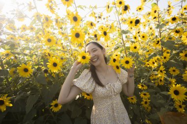 Mutlu genç Asyalı kadın yaz boyunca güzel ayçiçeği tarlasında neşeyle gülümsüyor. Giyinik neşeli bayan mükemmel bir günde doğanın ve parlak güneş ışığının tadını çıkarıyor.