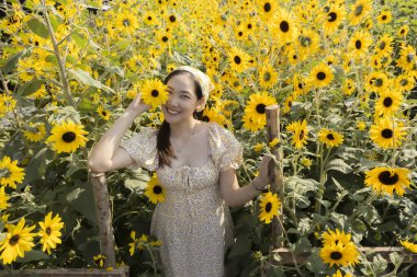 Güzel ayçiçeği tarlasında gülümseyen mutlu Asyalı kadın. Neşeli genç bayan güneşli bir günde sarı çiçek bahçesinde yaz doğasının tadını çıkarıyor. Güzelliğin neşeli portresi