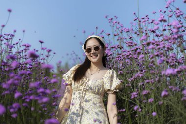 Mutlu gülümseyen Asyalı kadın, güneş gözlükleriyle güneşli yaz gününün tadını çıkarıyor. Doğadaki güzel mor verbena çiçek tarlasında duran neşeli insan.