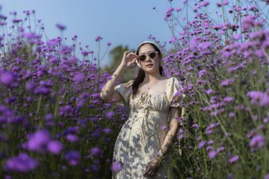 Şık bir elbise ve güneş gözlüğü takan mutlu genç Asyalı kadın güzel mor bir çiçek tarlasında dingin görünüyor, açık havada güneşli bir günün tadını çıkarıyor.