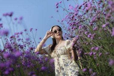 Mor verbena çiçek tarlasında kulaklıklı güzel Asyalı kadın müzik dinliyor. Mutlu ve kaygısız kız güneşli bir yaz günü doğada dinleniyor.