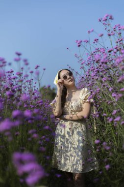 Mutlu Asyalı kadın mor çiçek tarlasında güzel bir yaz gününün tadını çıkarıyor. Bahçede genç bir yetişkin, doğada huzurlu bir ifadeyle berrak mavi gökyüzüne bakıyor.