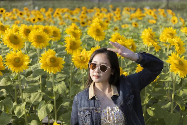 Güneş gözlüklü mutlu genç Asyalı kadın canlı sarı ayçiçeği tarlasında yaz yolculuğunun tadını çıkarıyor. Güzel doğada dinlenirken neşeli ve kaygısız bir duygu.