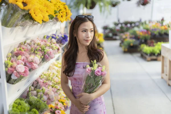Çiçekçinin içinde pembe elbiseli düşünceli genç Asyalı kadın elinde bir buket çiçek tutuyor. Markette durmuş, düşünceli bir ifadeyle başka tarafa bakıyor.