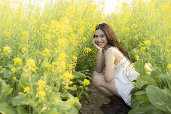 Güzel sarı çiçek tarlasında gülümseyen mutlu genç Asyalı kadın. Beyaz elbiseli hoş bayan, sakin bir yaz gününün tadını çıkarıyor.