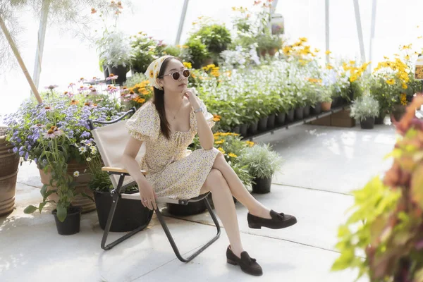 Güneşli serada sandalyesinde dinlenen sakin bir kadın. Bu yaşam tarzı onu güzel çiçek ve bitki düzenlemelerinin arasında otururken gösteriyor. Huzurlu ve mutlu hissediyor.