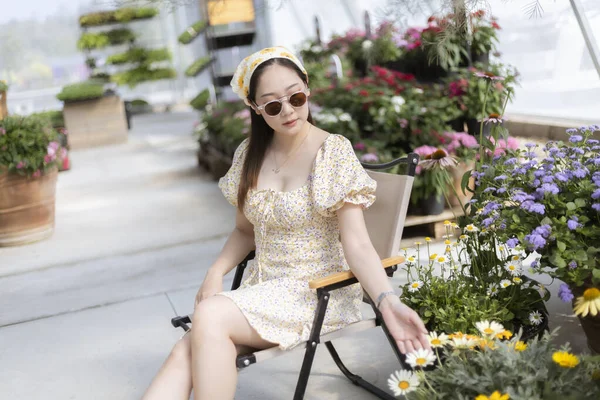 Güzel moda elbisesi ve beresi olan sakin genç Asyalı kadın serada sandalyede oturur, sakin atmosferin tadını çıkarır ve bahçede çiçeğe dokunur.