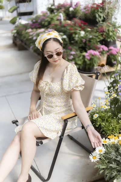 Sarı çiçekli ve güneş gözlüklü sakin Asyalı kadın bahçede sandalyede dinleniyor. Beyaz çiçeğe hafifçe dokunur ve dışarıda güneşin tadını çıkarır.