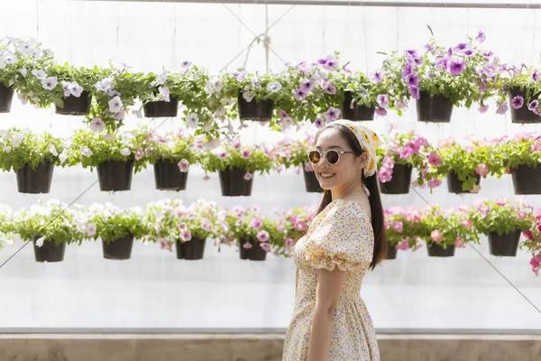 Bahçede neşeyle gülümseyen güneş gözlüklü mutlu Asyalı kadın. Arka planda bahar mevsiminde serada asılı petunya saksıları duvarı yer alıyor