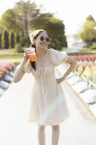 Yazlık elbiseler içinde gülümseyen mutlu genç bir kadın. Parkta güzel bir günün tadını çıkarırken elinde içki tutuyor. Güneş gözlüklü neşeli yaşam tarzı konsepti