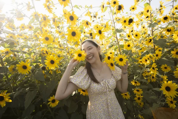 Mutlu genç Asyalı kadın yaz boyunca güzel ayçiçeği tarlasında neşeyle gülümsüyor. Giyinik neşeli bayan mükemmel bir günde doğanın ve parlak güneş ışığının tadını çıkarıyor.