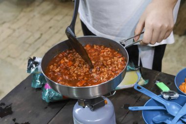 Odaklanmış aşçı tuzlu bolonez sosunu kıyma ve tavada domatesle karıştırıyor. Lezzetli ev yapımı kamp yemeği için portatif gaz ocağında açık hava yemeği hazırlığı.