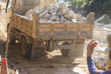 İnşaat yolunda ağır bir çöp kamyonu var. Taş ve kaya taşıyorlar. İşçi endüstriyel ekipman yönetiyor, yoğun iş gücü ve toz sahnesi