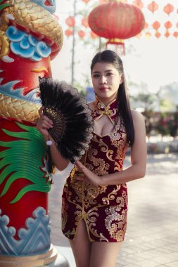 Zarif, kırmızı, geleneksel Çin Cheongsam elbisesi giymiş kendine güvenen Asyalı kadın yelpaze tutuyor. Çin Yeni Yıl kutlamaları için açık hava tapınağında güzel bir portre.