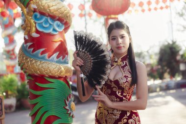 Kırmızı geleneksel Çin peyniri giymiş zarif Asyalı kadın elinde yelpaze tutuyor. Tapınaktaki renkli ejderha sütununun yanında duruyor.