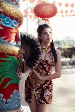 Siyah yelpaze tutan geleneksel kırmızı Çin peyniri elbiseli zarif Asyalı kadın. Zarif bayan model, yeni yılı kutlamak için tapınakta kırmızı fenerlerle poz veriyor.
