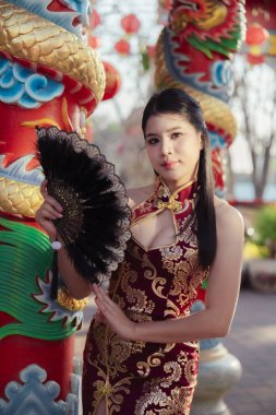 Geleneksel kırmızı Çin Qipao elbisesi içinde güzel bir Asyalı kadın siyah yelpaze tutuyor, kültürel ejderha sütunlarına karşı sakin ve zarif bir görünüm sergiliyor.