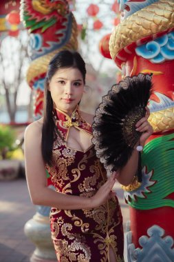 Geleneksel kırmızı Cheongsam elbisesi içindeki kendine güvenen güzel Asyalı kadın siyah yelpaze tutuyor. Çin kültürünü ve açık havada renkli ejderha heykelinin yanında modayı kutluyor.