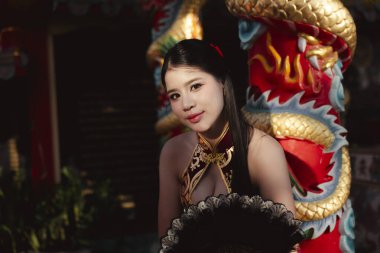 Geleneksel siyah Cheongsam elbisesi içinde kendine güvenen genç Asyalı kadın, sıcak akşam ışığında çekici bir bakışla renkli Çin ejderha heykelinin yanında duruyor.