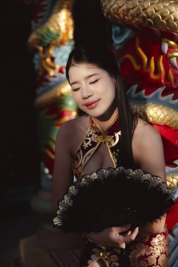 Geleneksel Çinli elbiseli sakin genç Asyalı kadın siyah yelpaze tutuyor. Tapınağın içinde altın ejderha heykelinin yanında oturuyor. Güzel yüzü yumuşak ışıkta sakin.