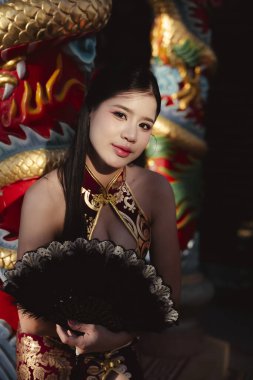 Geleneksel fantezi kostümlü zarif bir Asyalı kadının kendine güvenen portresi elinde siyah yelpaze, güzellik çekimi için renkli Çin ejderhası arka planına karşı poz veriyor.