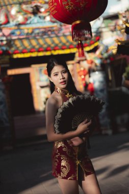 Geleneksel kırmızı Çin Cheongsam elbisesi giymiş kendine güvenen genç Asyalı kadın yelpaze tutuyor. Kültürel kutlama sırasında Çin Mahallesi 'nde kırmızı fenerle yapılan zarif bir portre.