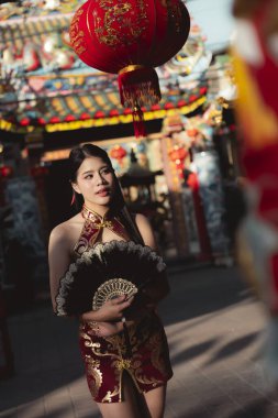Kırmızı Cheongsam elbiseli düşünceli Asyalı kadın Çin Yeni Yılı 'nı kutluyor. Canlı kırmızı fener dekorasyonunun altında zarif bir şekilde elinde yelpaze tutuyor.