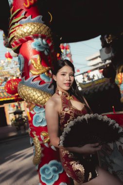 Geleneksel kırmızı elbiseli güzel Asyalı kadın Çin Yeni Yıl kutlamalarında mutluydu. Tapınakta ejderha sütununun yanında duruyor, kendine güvenen bir bakışla yelpaze tutuyor.