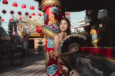 Geleneksel Cheongsam 'daki dingin genç Asyalı kadın Çin Yeni Yıl Festivali' nde hayranı, tapınak avlusunda kırmızı fenerlerle aslan dansı kostümünün yanında duruyor.