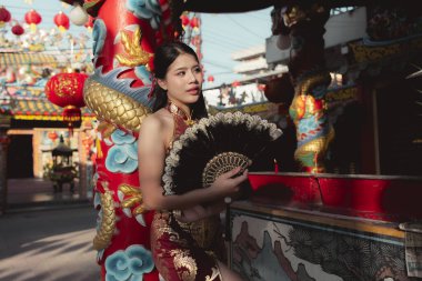 Geleneksel Çin elbisesi giymiş sakin genç Asyalı kadın tapınakta yelpaze tutuyor. Onun düşünceli ifadesi kültürü ve coşkulu festivalin kutlamasını yansıtıyor.